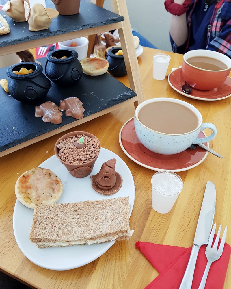 A photo of Sam's plate at Harry Potter afternoon tea. On the plate is a mini cheese pizza, chocolate fudge in the shape of a sorting hat, a chocolate mandrake pot and a dainty sandwich. Next to her plate is a cup of coffee and a shot glass filled with "butterbeer"