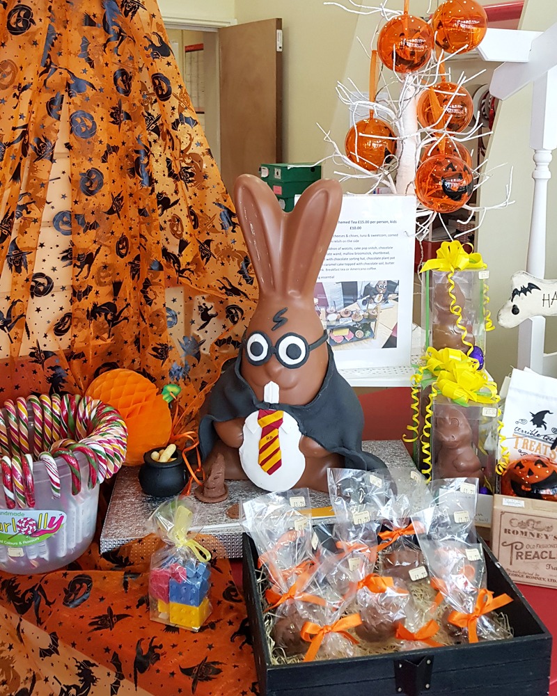 A chocolate-shaped bunny dressed like Harry Potter is sat on a display table, surrounded by Halloween decorations. The bunny has a lightening bolt scar, big glasses and is wearing a black cape with a red and yellow stripey tie