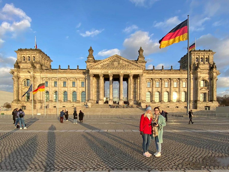 Sam and Jess are posing for a photo in front of the Reichstag building. The German flag is blowing in the wind in front of a clear blue sky.