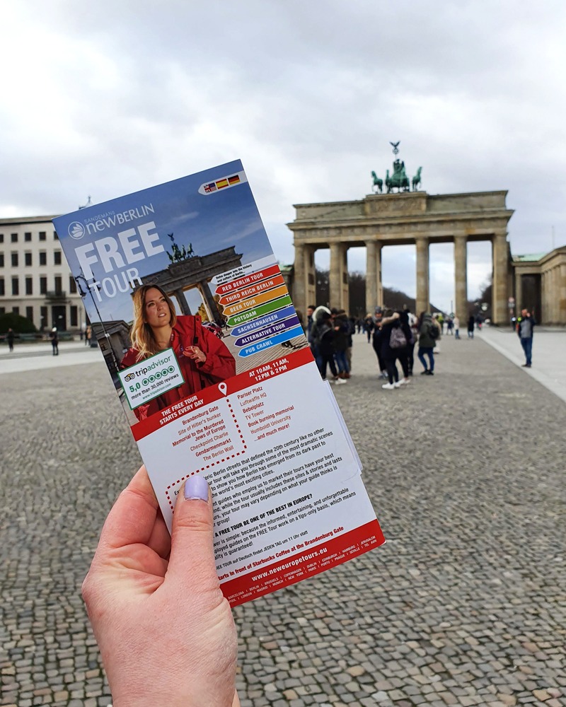 Sam is holding up a leaflet in front of the Brandenburg Gate. It reads "Free Walking Tour - rated 5 on TripAdvisor"