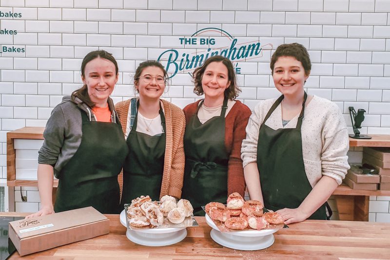 Sam and her family are wearing aprons, smiling at the camera while standing in front of a sign which says "The Big Birmingham Bake"
